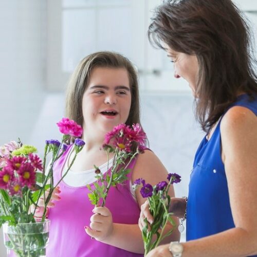 A teenage girl with down syndrome arranging flowers with her mother. They are holding cut flowers in their hands, looking at each other, smiling and talking.